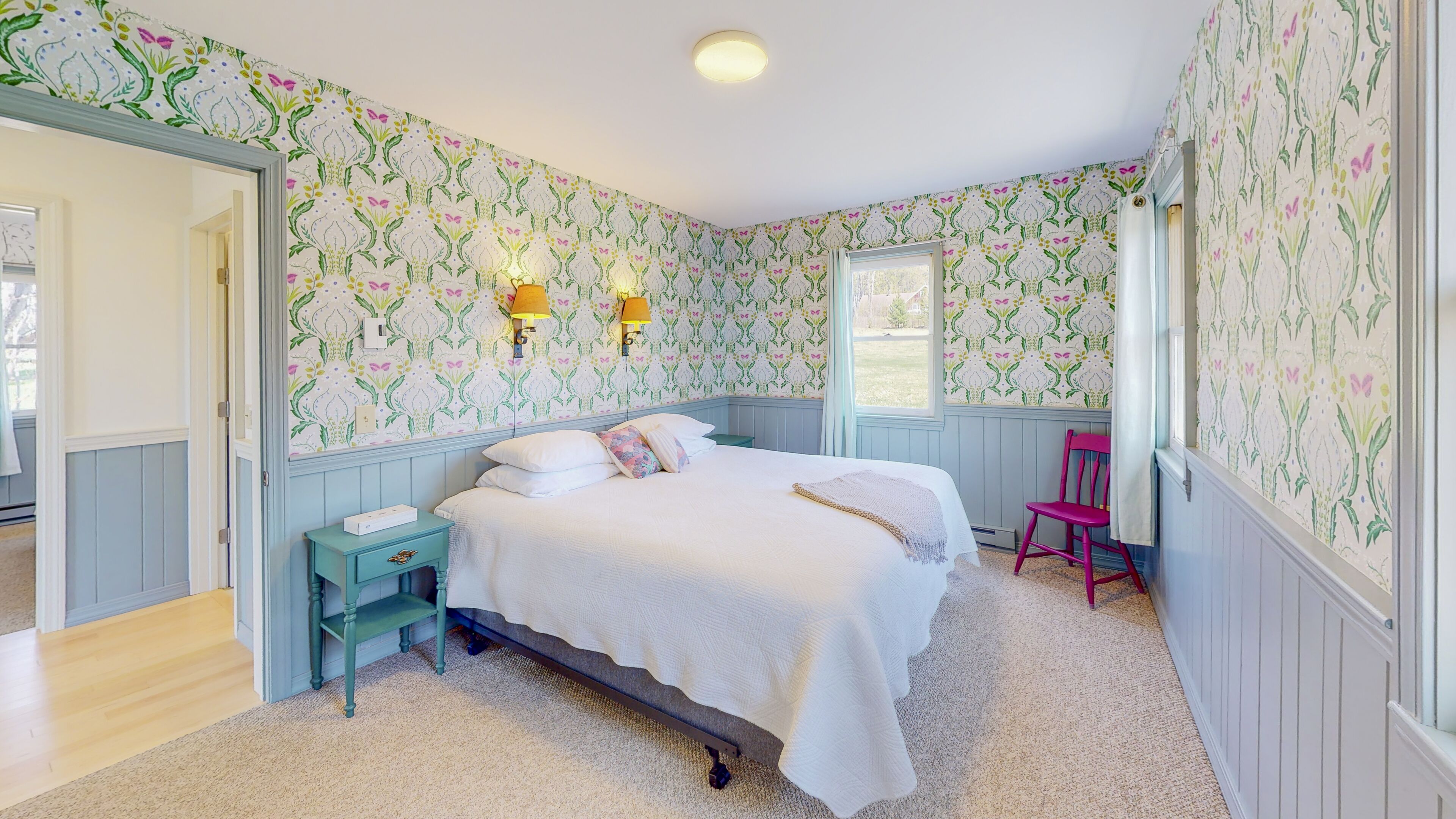 Une chambre à coucher avec un papier peint à motifs floraux, un lit recouvert de draps blancs, une table de chevet, une chaise et une fenêtre donnant sur un beau paysage.