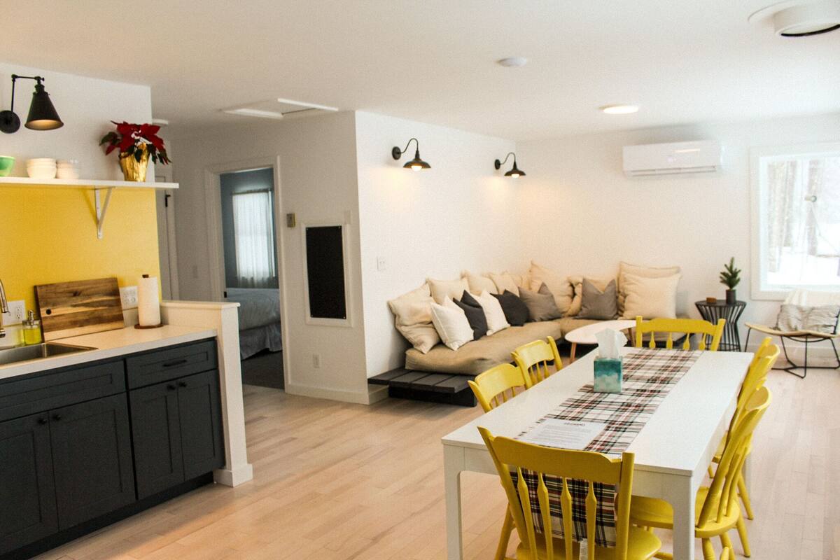 A modern kitchen with a dining area, featuring a white table, yellow chairs, and a sofa with multiple cushions.