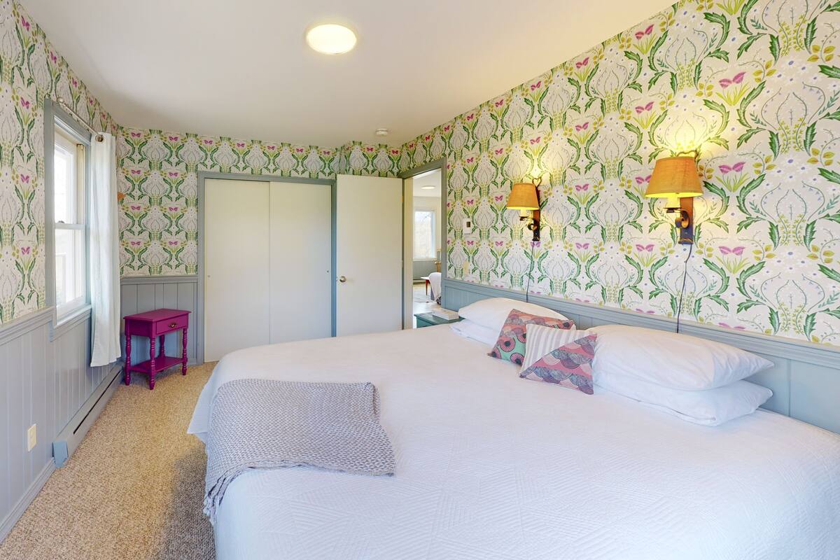A bedroom with floral wallpaper, a bed with white linens, two bedside lamps, and a small pink nightstand.