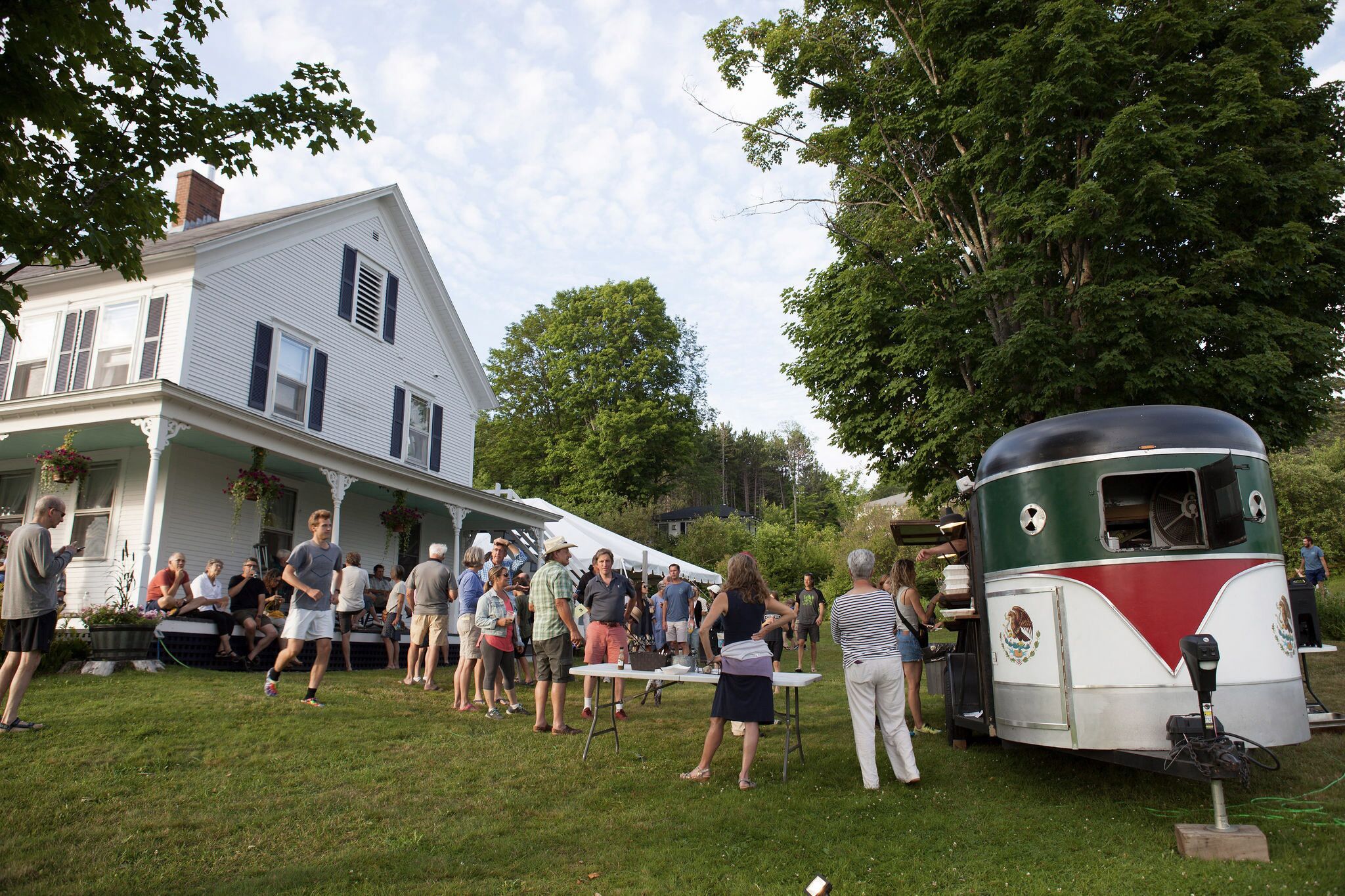 A group of people gathered outdoors near a house and a vintage trailer.