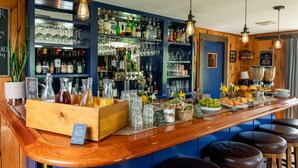Een bar met een houten toog, krukken en planken vol flessen en glazen.