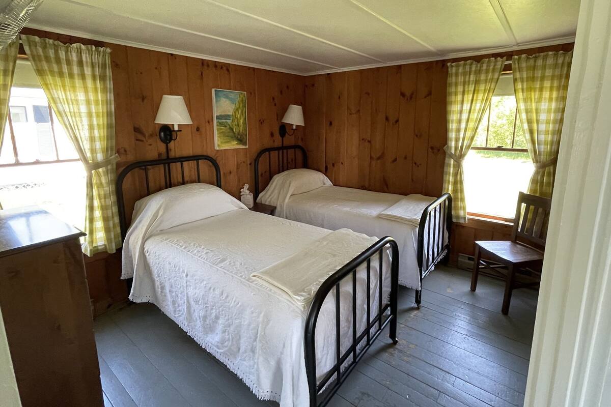A room with two beds, wooden walls, a window with curtains, and a table with a chair.