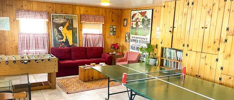 A room with a foosball table, ping pong table, red sofa, and wooden walls.