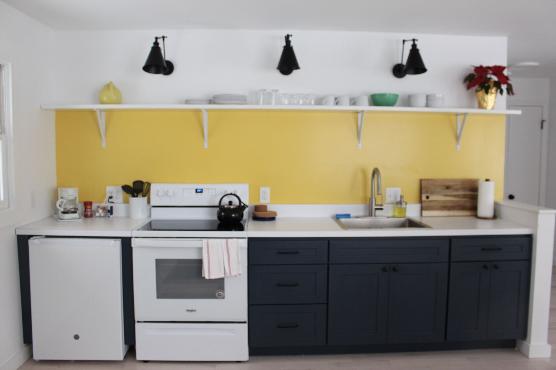 Une cuisine moderne avec un four blanc, un évier et des meubles de couleur sombre. Des luminaires sont suspendus au-dessus du plan de travail, et un pare-feu jaune décore le mur.