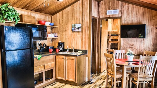 A compact kitchen with wooden cabinets, a black refrigerator, a microwave, and a dining area with wooden chairs and a table. - American Heritage RV Park (Williamsburg)
