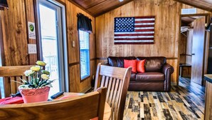 A cabin interior with a wooden floor, a brown leather sofa, a table with a pink pot of flowers, and an American flag on the wall. - American Heritage RV Park (Williamsburg)