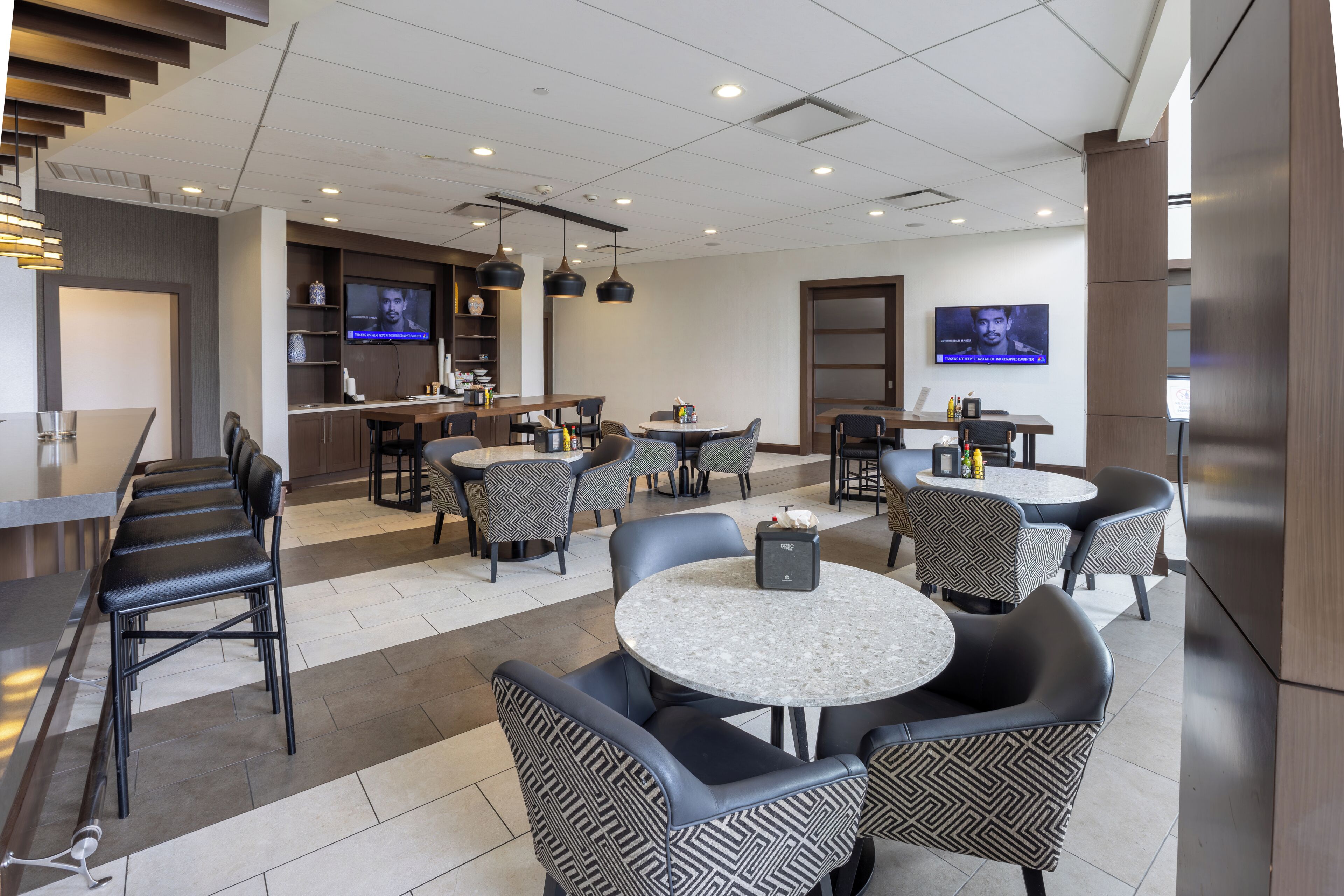 Un moderno vestíbulo de hotel con varias zonas de estar, incluyendo taburetes de bar y mesas redondas, y un televisor en la pared.