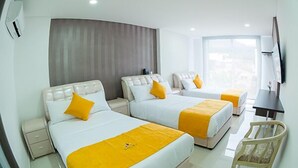 A hotel room with three beds, each with a white headboard and yellow accents, a desk with a chair, and a large window providing a city view. - Solev Hotel (Villavicencio)