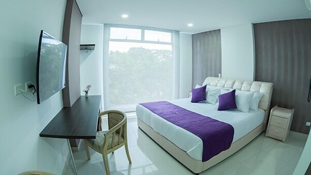A modern hotel room with a large bed, a desk, a chair, a television, and a window with a view of trees. - Solev Hotel (Villavicencio)