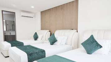 Une chambre d’hôtel avec trois lits, chacun doté de draps blancs et de coussins décoratifs verts. On y trouve une tête de lit en bois, un mur blanc et un appareil de climatisation fixé au mur.