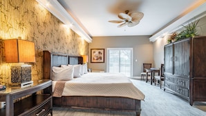 Une chambre à coucher avec un grand lit, une table de chevet, une commode, un ventilateur de plafond et une fenêtre avec des stores.