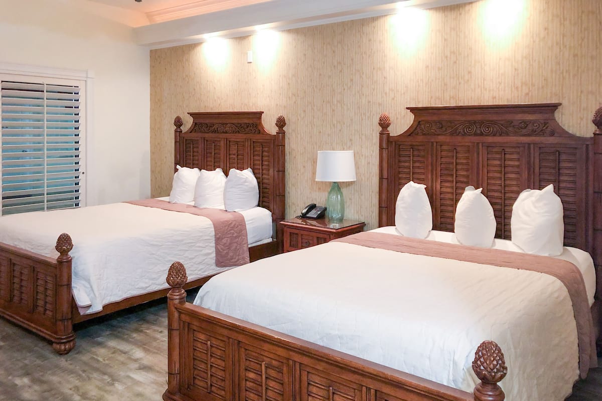Una habitación de hotel con dos camas de madera, una mesita de noche con lámpara y una ventana con persianas.