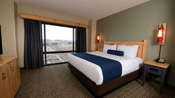 A hotel room with a large bed, a wooden headboard, a nightstand, a lamp, and a view of a cityscape through a large window.