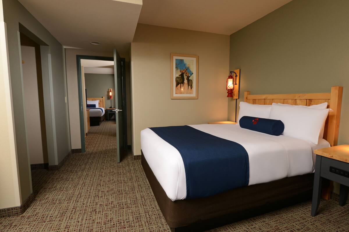A hotel room with a large bed, a wooden headboard, a nightstand, a wall-mounted lamp, and a painting of deer.