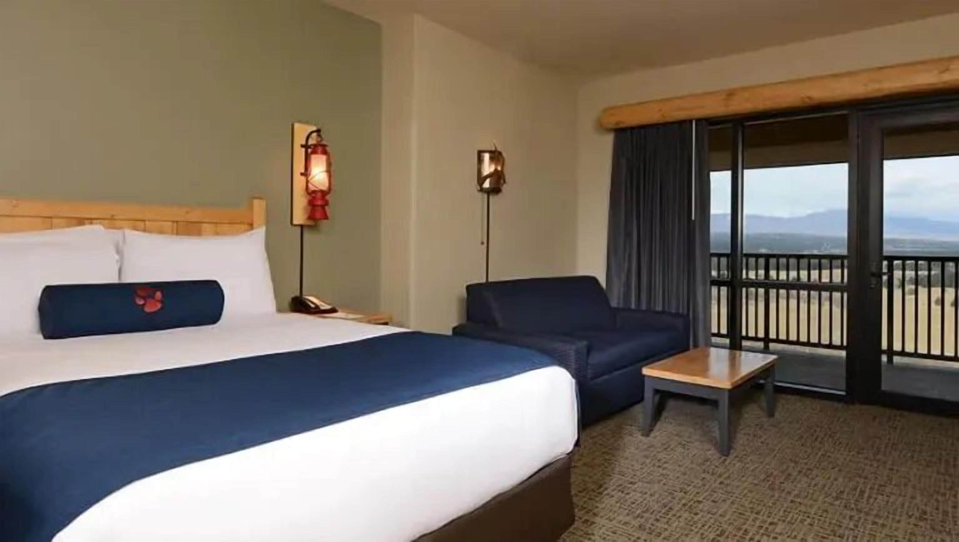A hotel room with a bed, a sofa, a small table, a nightstand, and a balcony with a view.