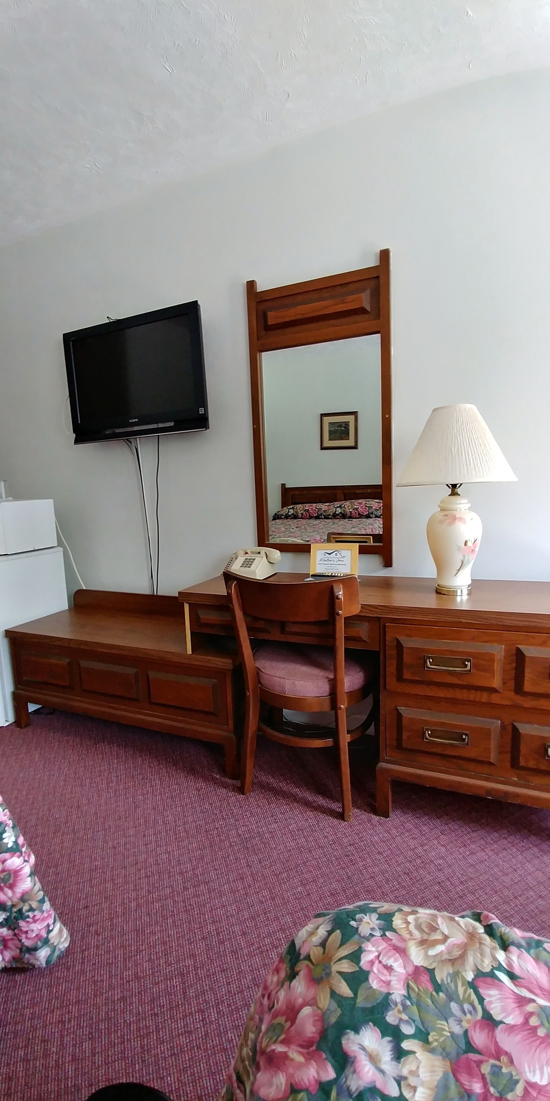Una scrivania in legno con cassetti, una sedia, un telefono, una lampada e un televisore fissato alla parete.