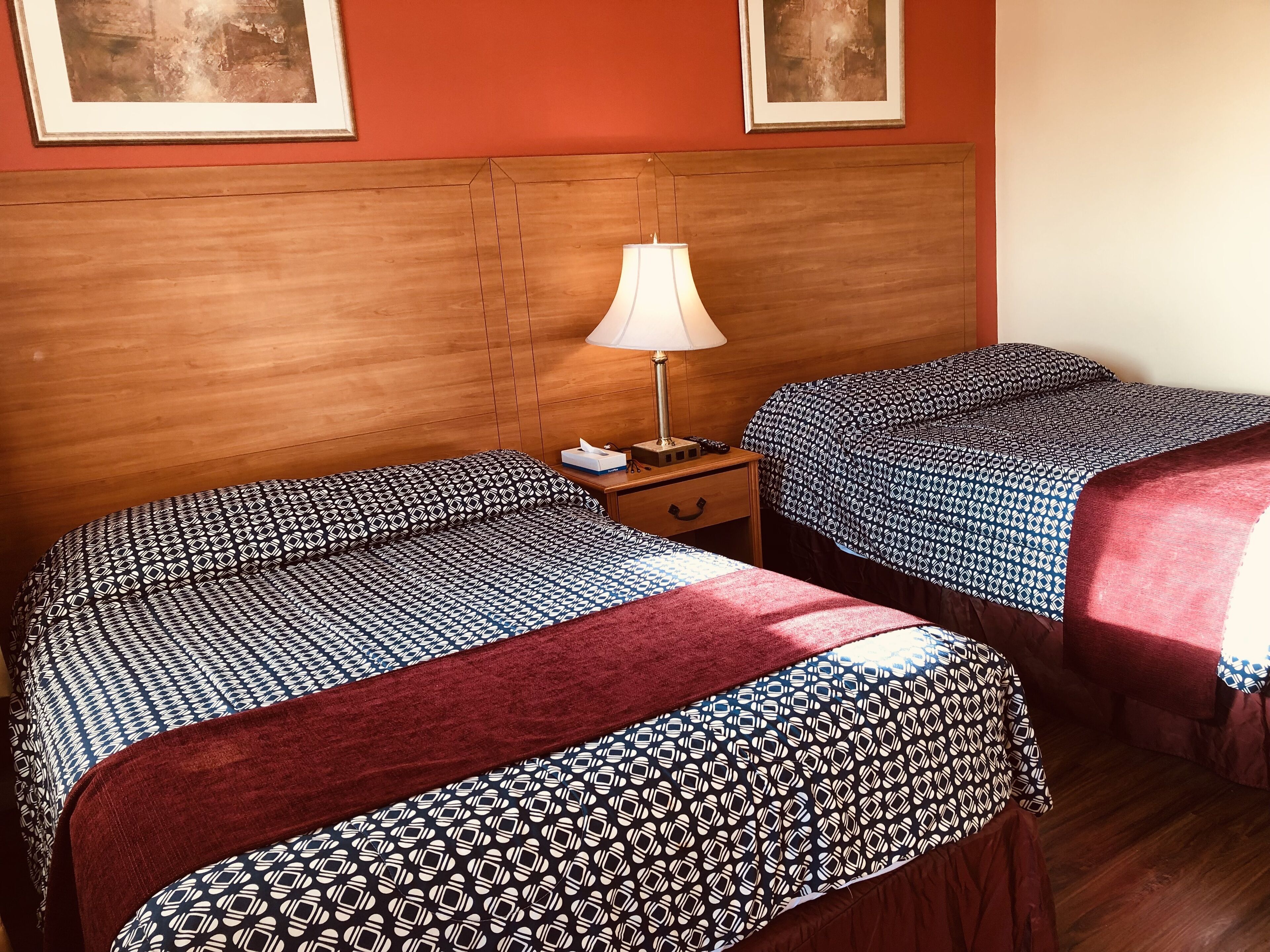 Habitación de hotel con dos camas, cabecero de madera, una mesita de noche con lámpara y dos cuadros enmarcados en la pared.