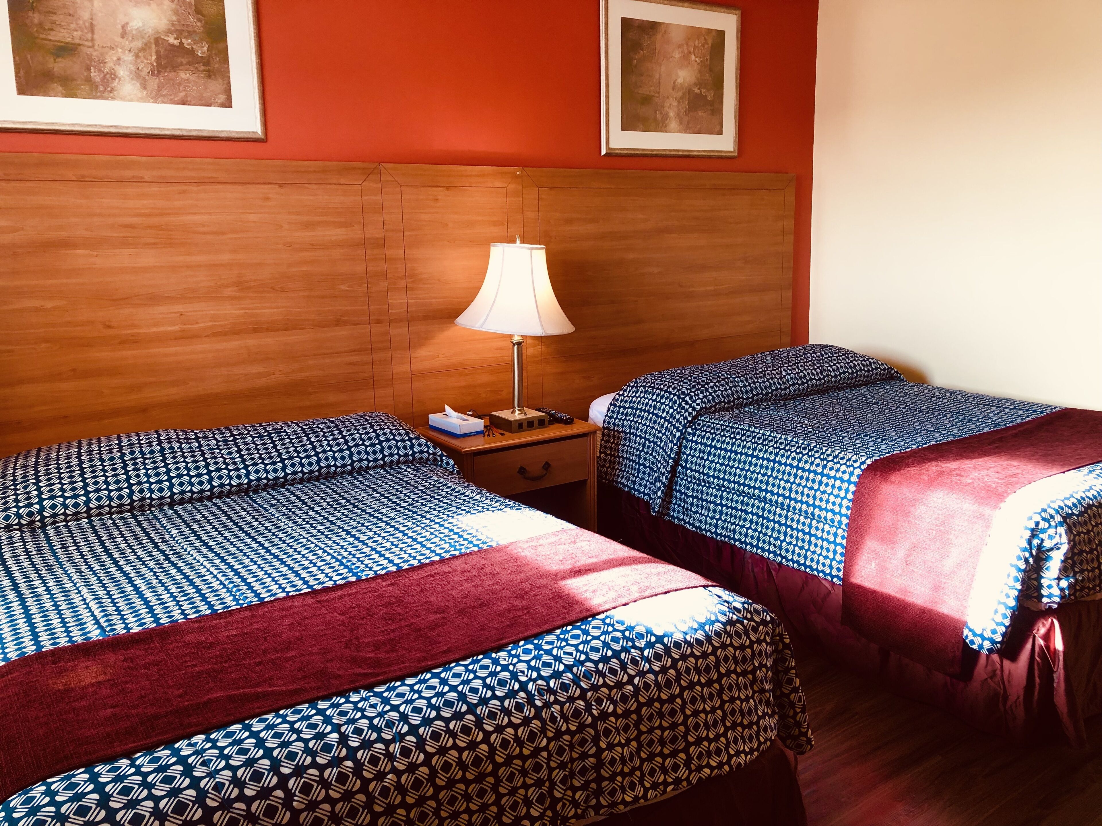 Quarto de hotel com duas camas, cabeceira de madeira, mesa de cabeceira com abajur e quadros nas paredes.