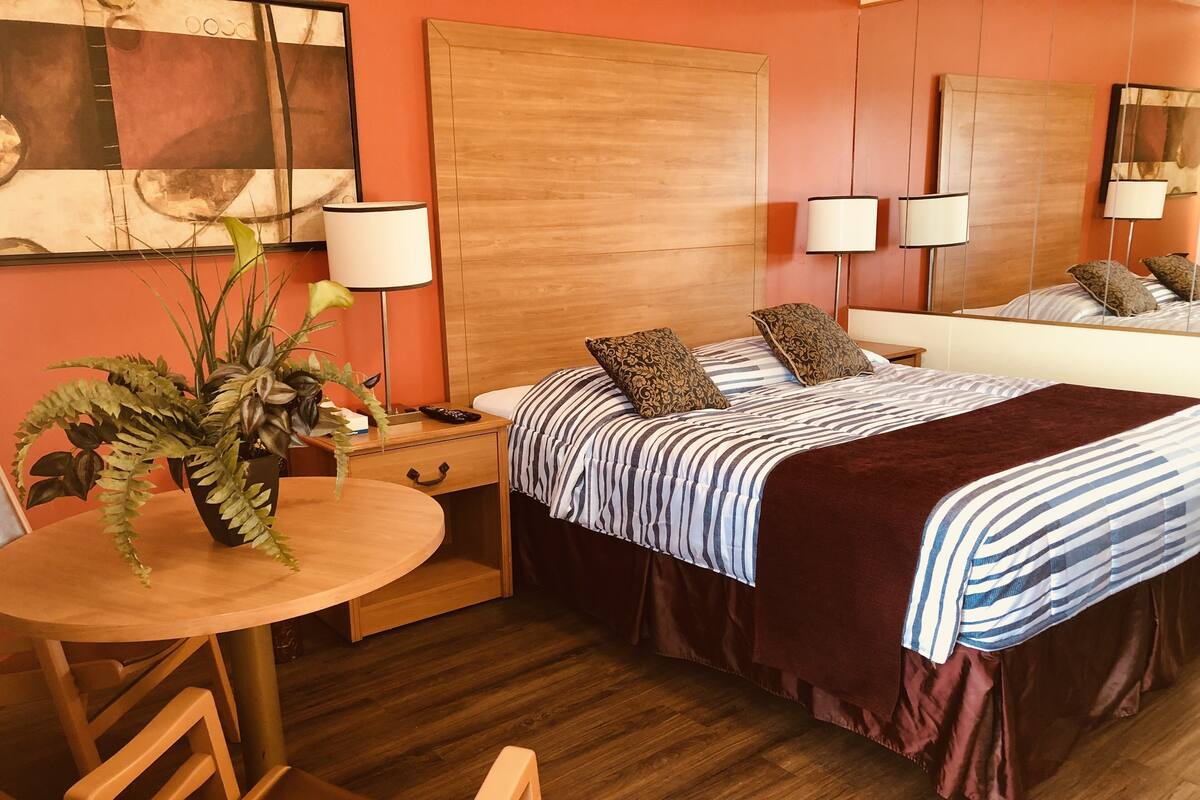 A hotel room with a wooden headboard, a bed with striped bedding, two bedside lamps, a small table with a plant, and a chair.