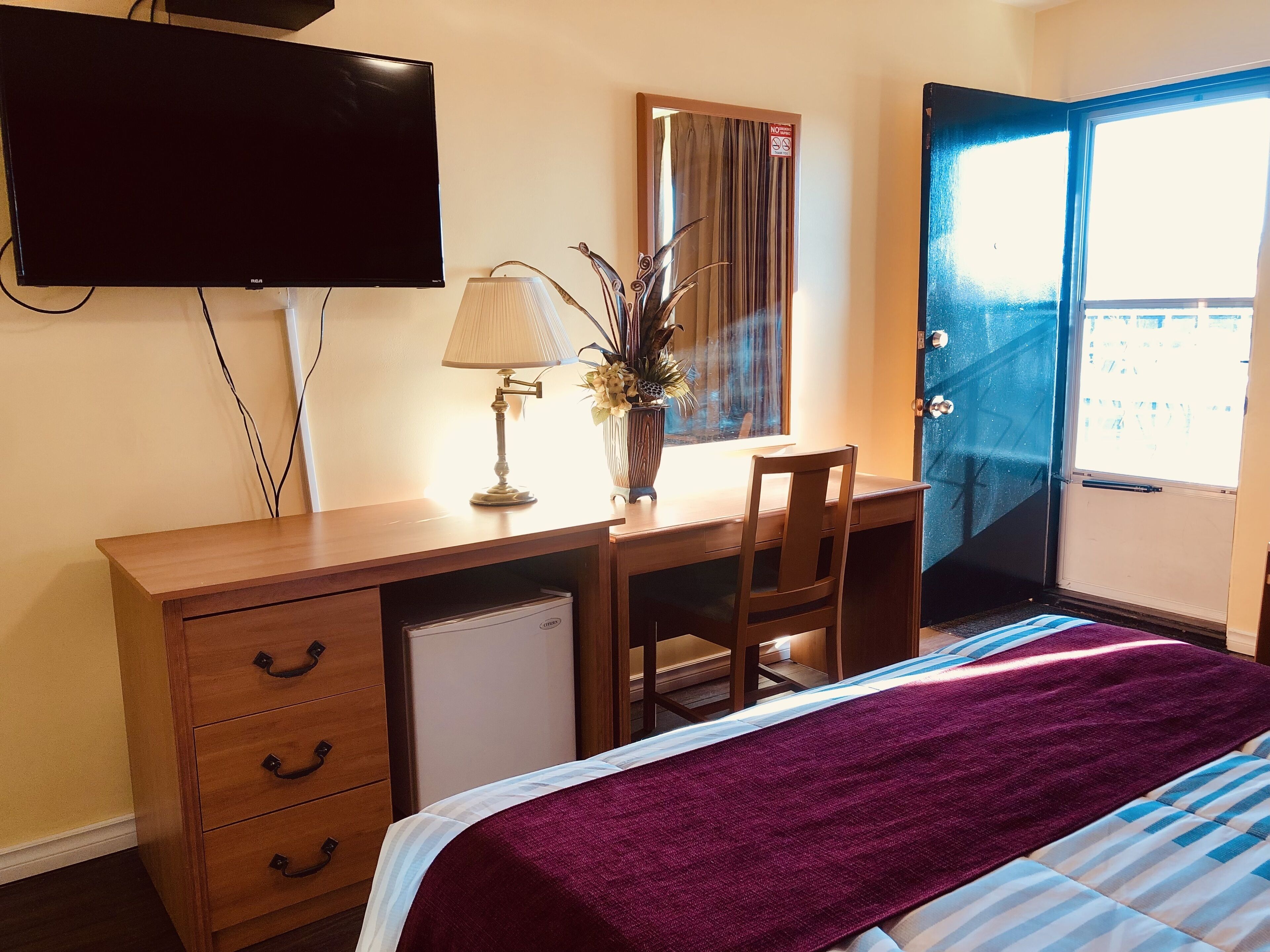 A hotel room with a wooden desk, a television, a lamp, a chair, a bed with a purple blanket, and a window with a view.