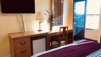 A hotel room with a wooden desk, a television, a lamp, a chair, a bed with a purple blanket, and a window with a view.