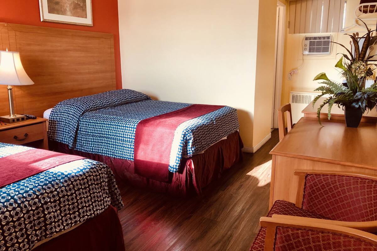 A hotel room with two beds, a wooden headboard, a desk with a chair, a potted plant, and a window with blinds.