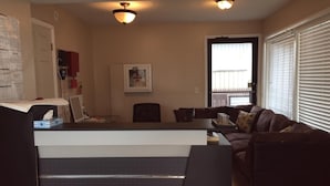 Ein Wohnzimmer mit einer Couch, einem Couchtisch und einem Schreibtisch mit Stuhl. Es gibt ein Fenster mit Jalousien, eine Tür und eine Deckenleuchte.