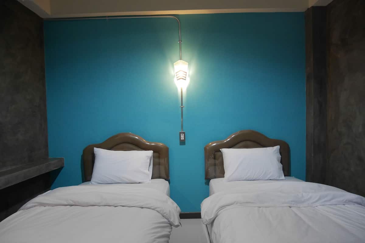 A room with two beds, each with a wooden headboard and white bedding. A wall-mounted light fixture is visible above the beds.