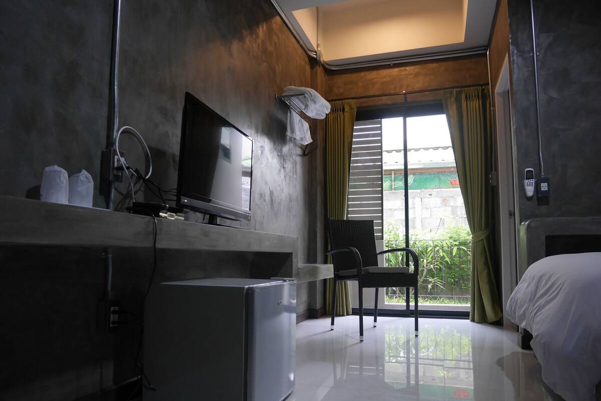 A hotel room with a bed, a TV, a small refrigerator, a chair, and a window with curtains.