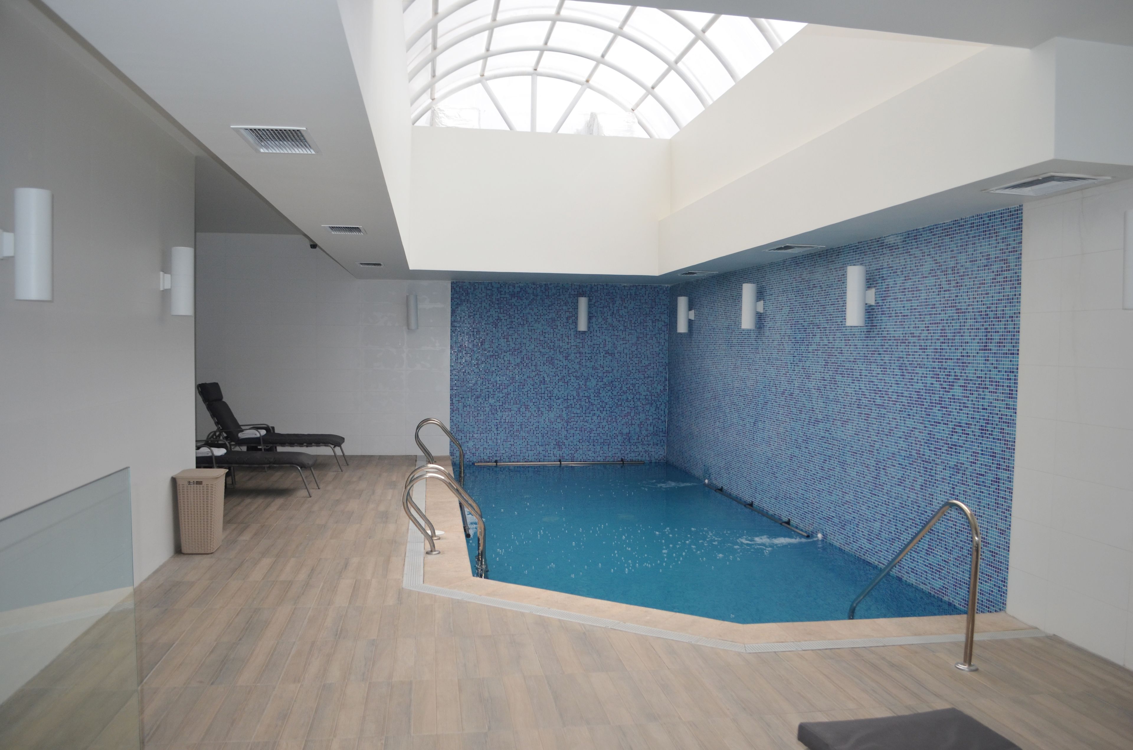 An indoor swimming pool with a skylight, a lounge area with a chair, and a tiled wall.