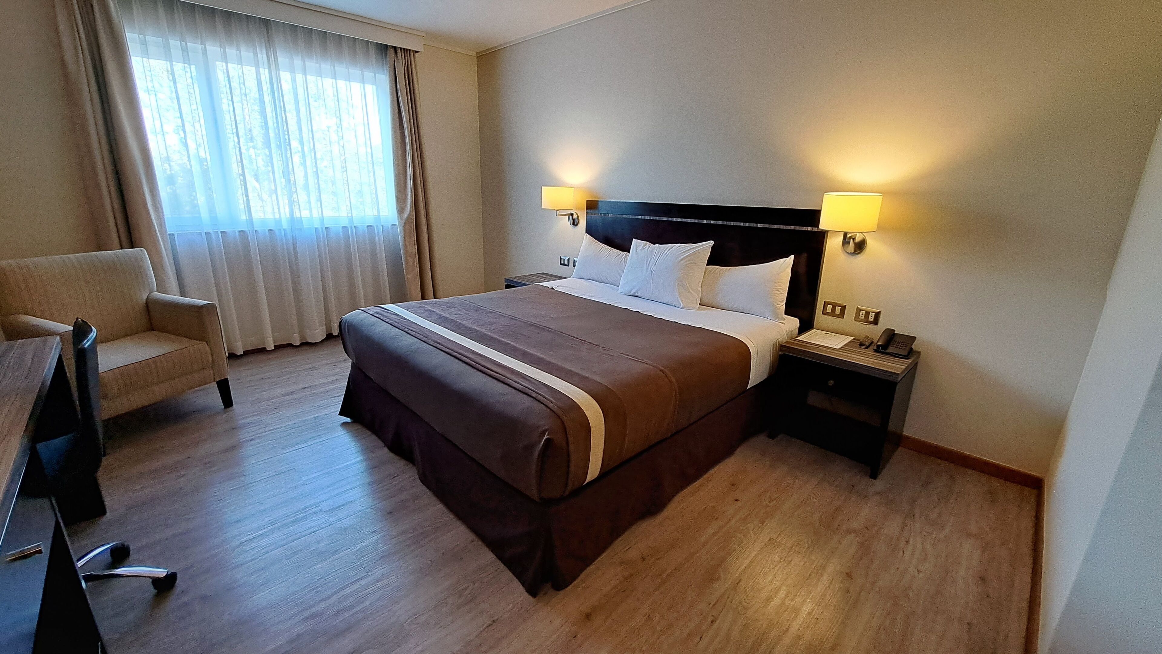 A hotel room with a large bed, a desk, a chair, and a nightstand with a phone and lamp.