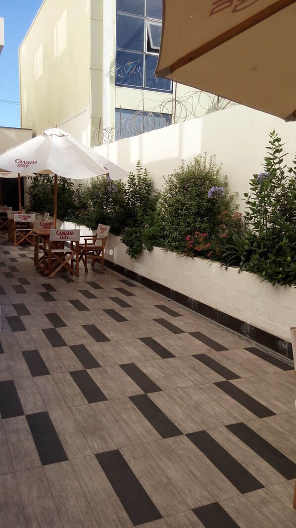 An outdoor seating area with tables and chairs, some with 'Canada Dry' signs, and a white wall with barbed wire.