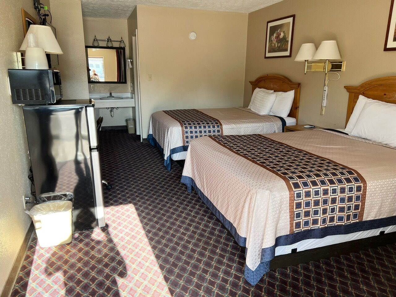 A hotel room with two beds, a desk with a lamp, a mirror, and a small trash bin.