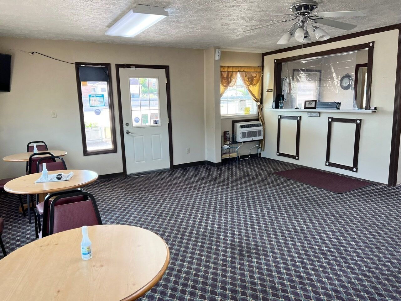 A room with two doors, a ceiling fan, a window with curtains, a wall-mounted air conditioner, and a welcome sign.