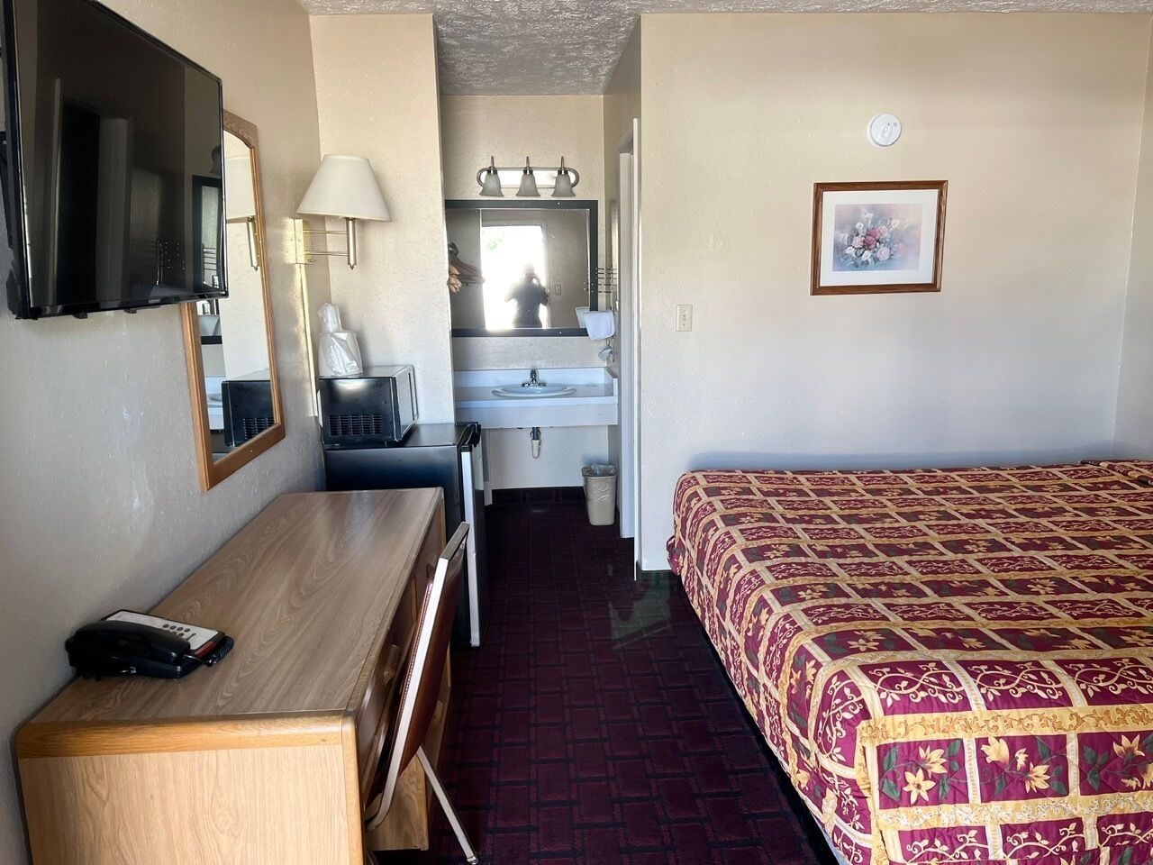 Une chambre d’hôtel avec un bureau en bois, un lit recouvert d’une courtepointe à motifs, un miroir, un lavabo et un téléviseur.