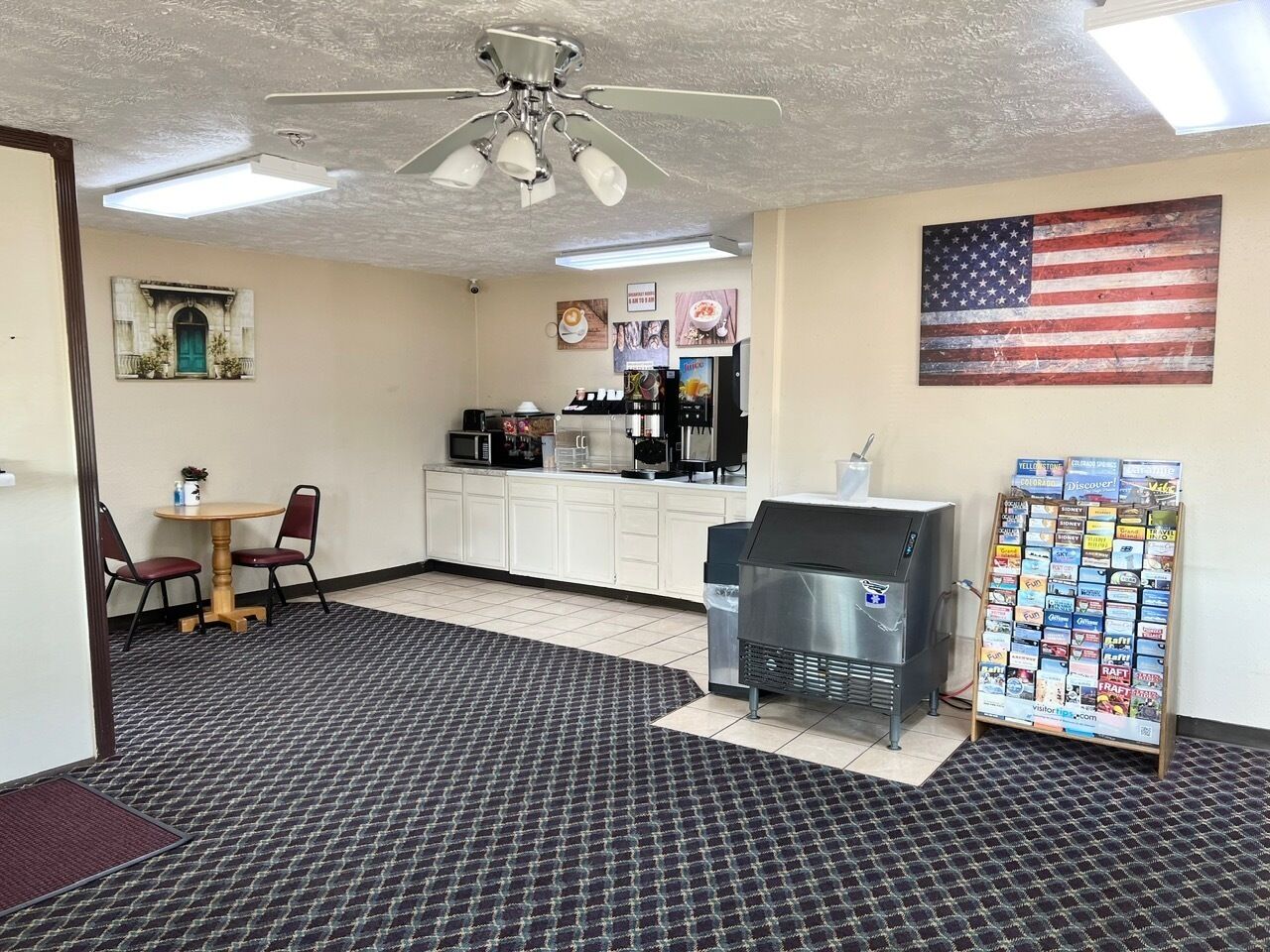 A room with a kitchenette, a ceiling fan, a coffee machine, a grill, and a bulletin board with travel brochures.
