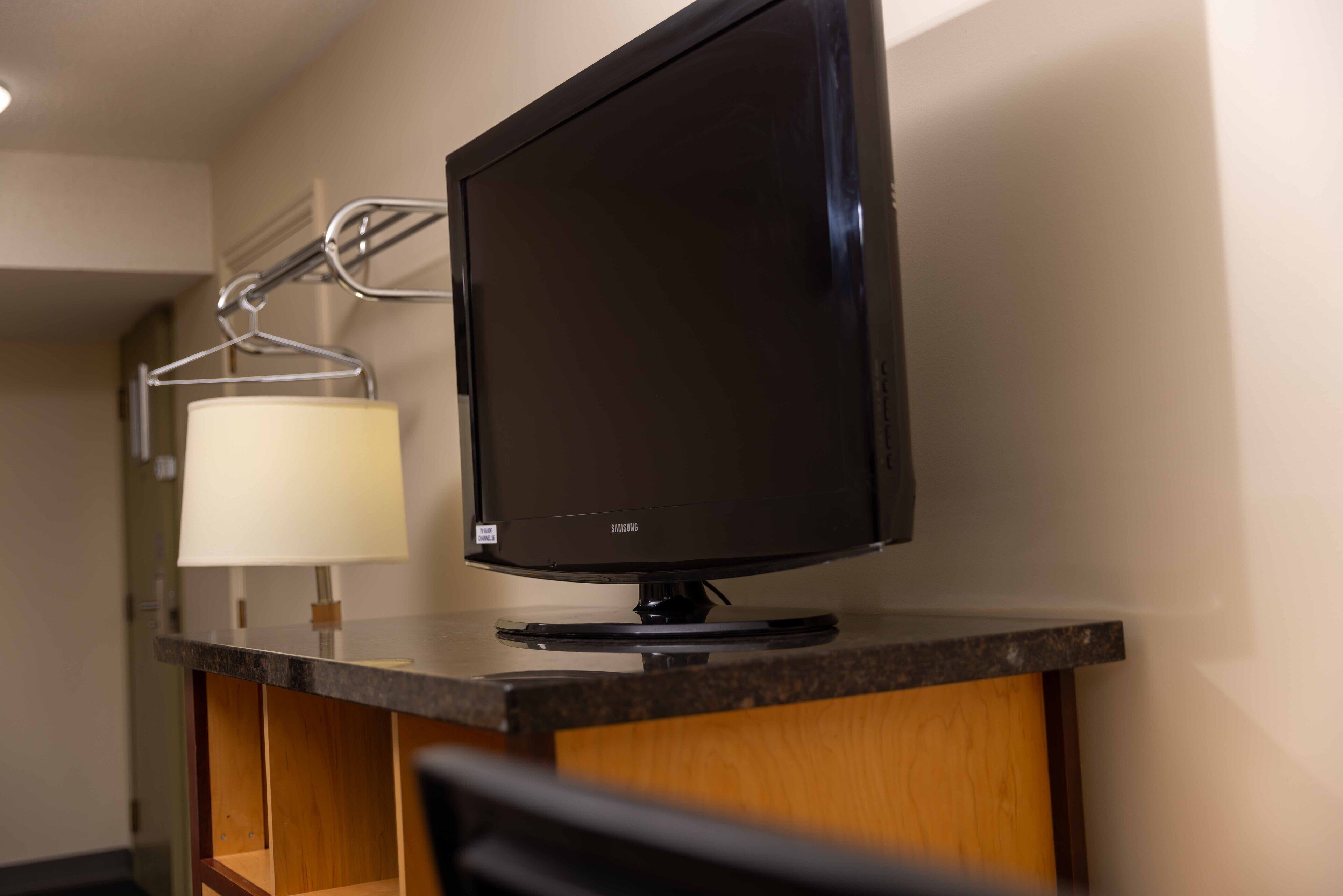 A hotel room with a flat-screen TV on a wooden desk, a lamp, and a wall-mounted rack.