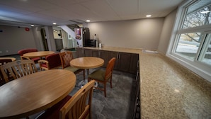 Un comedor limpio y moderno con mesas y sillas de madera, una encimera de cocina y un amplio ventanal con vistas al exterior.