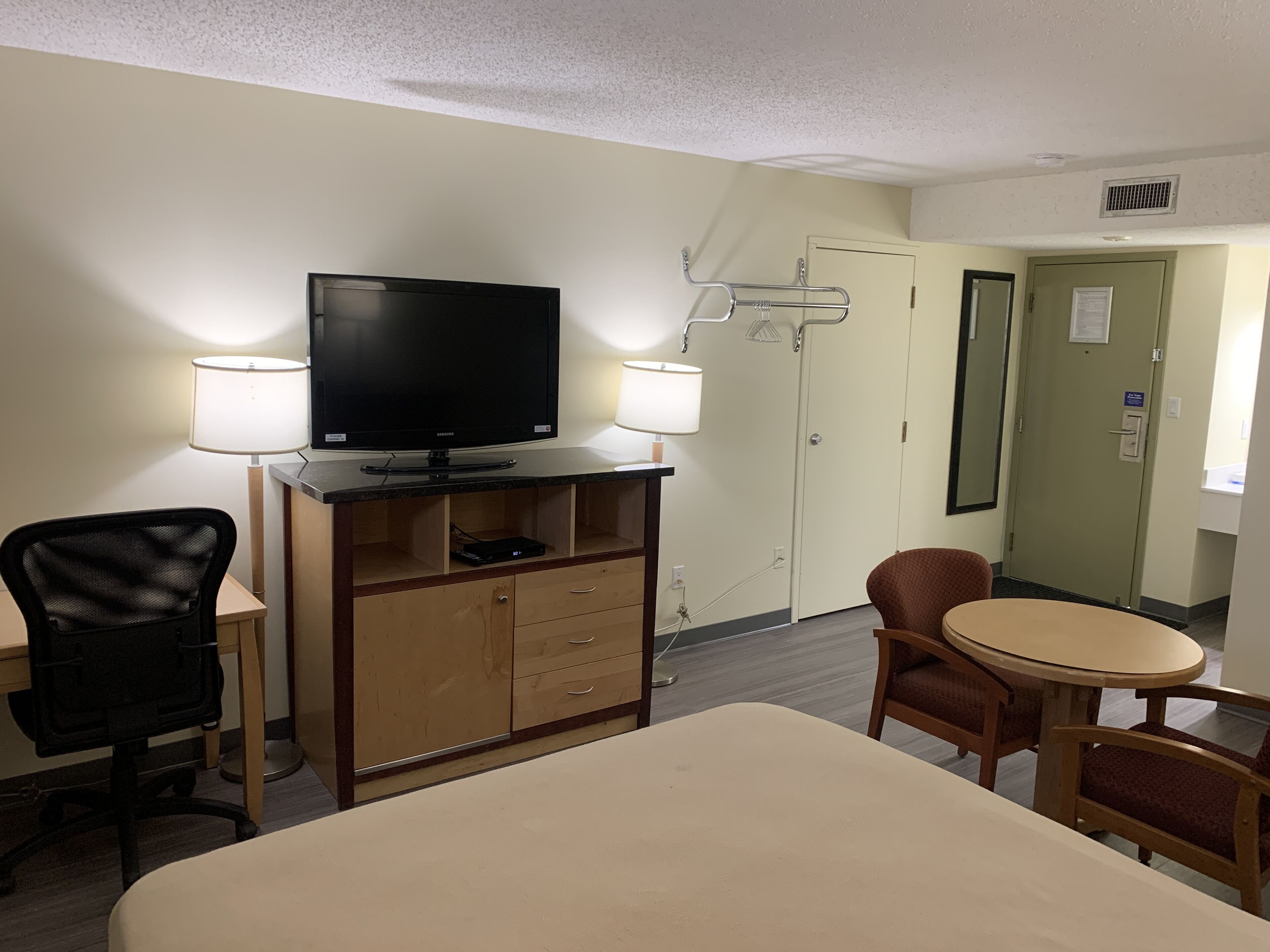 A hotel room with a bed, a TV, a desk with a lamp, a chair, and a small table.