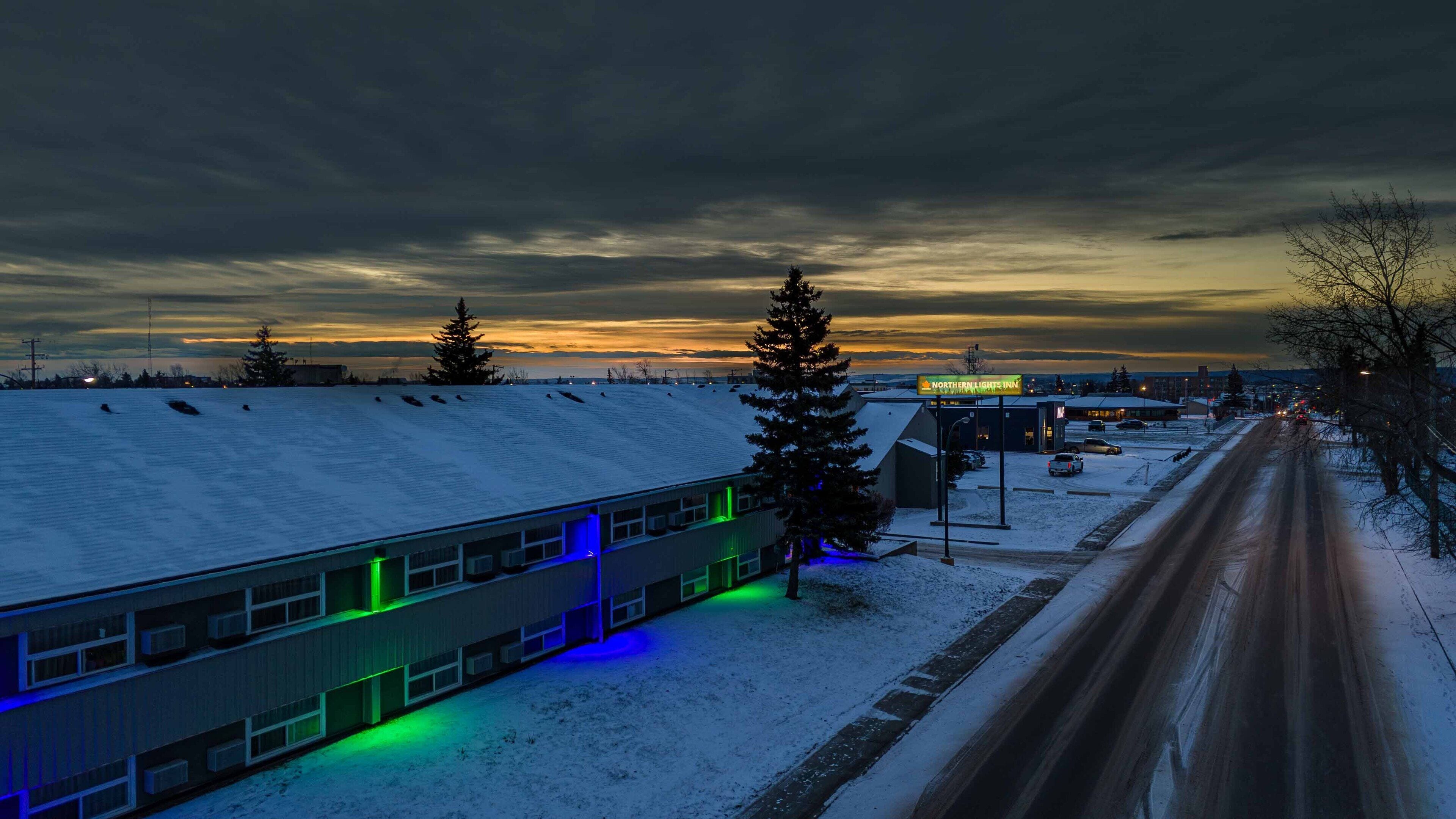 Paisaje nevado con un edificio que tiene ventanas iluminadas en verde y azul, un letrero que dice Northern Lights Inn, y una carretera con vehículos.