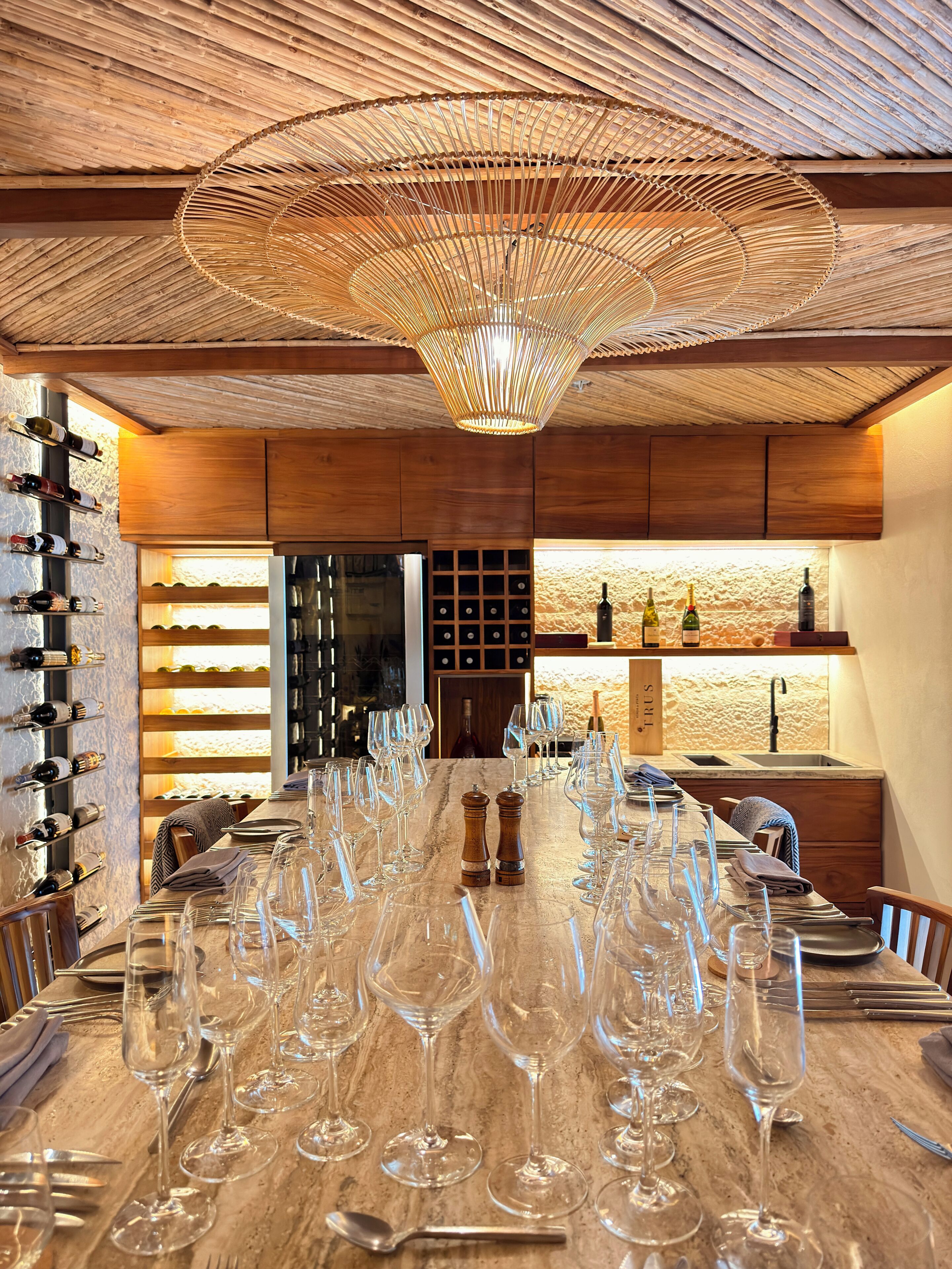 A dining area with a long table set for a meal, wine bottles on shelves, and a unique ceiling design.