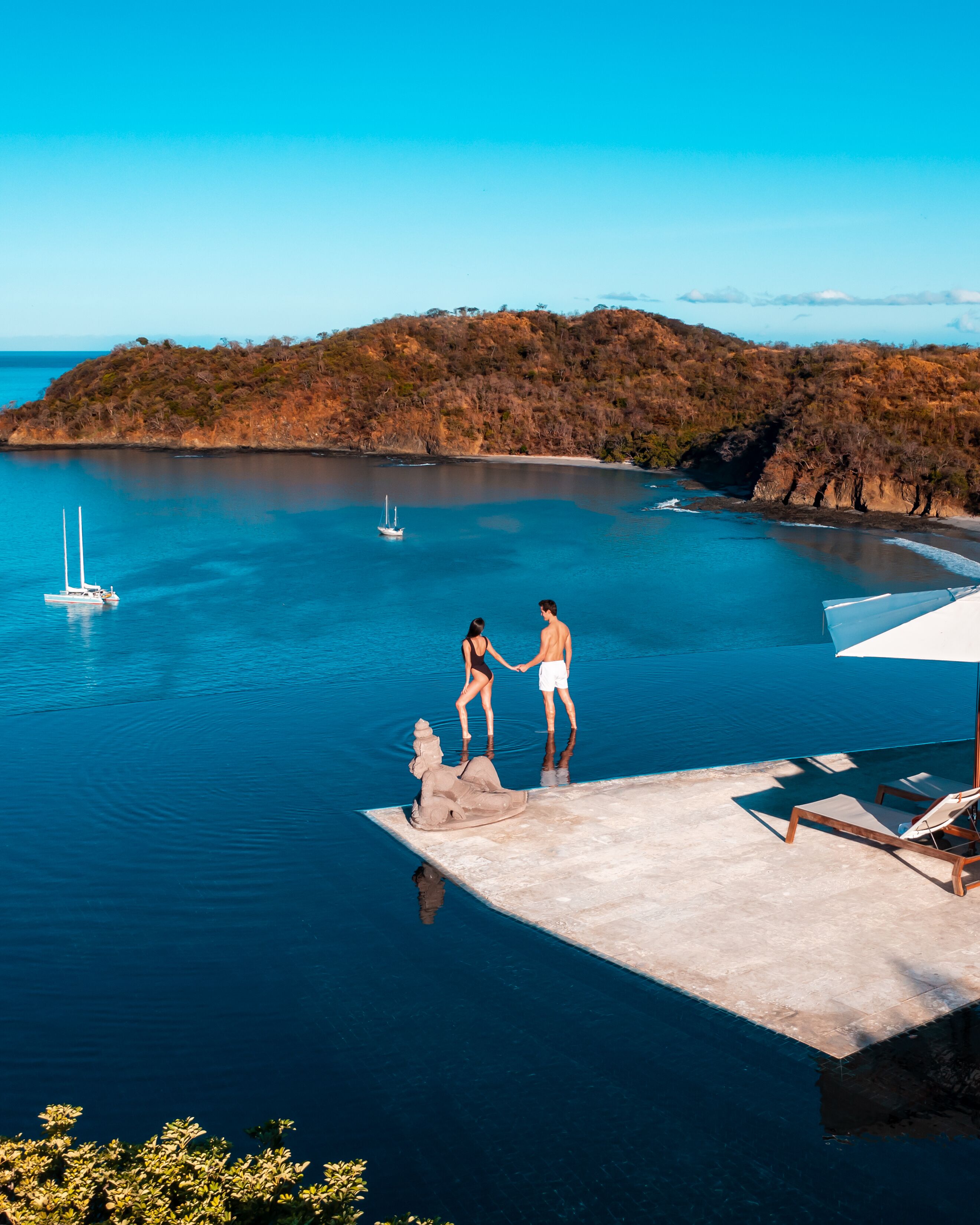 Una pareja está de pie en la terraza de una piscina, con vista a un mar tranquilo y una isla distante.