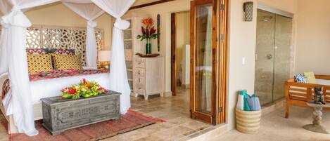 Une chambre à coucher avec un lit à baldaquin, une table de chevet ornée d’un vase de fleurs et un banc en bois surmonté d’une statue.
