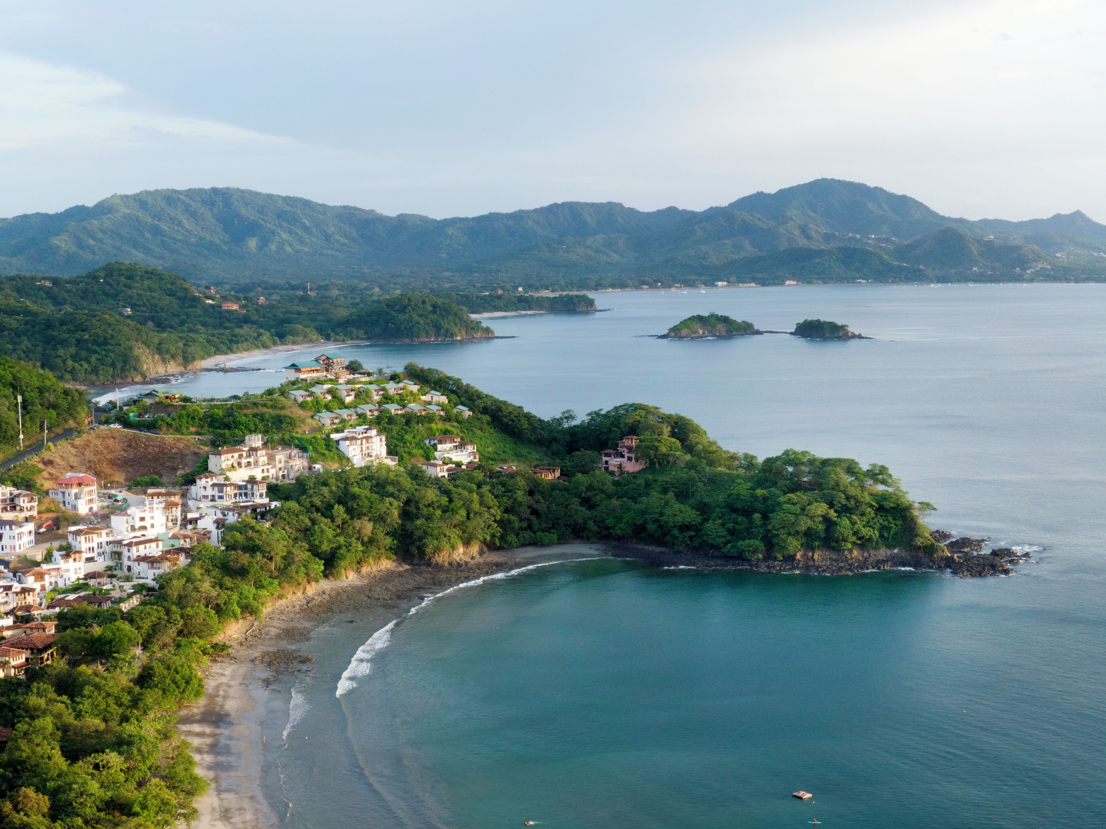 An aerial view of a coastal town with buildings along the shoreline, surrounded by lush greenery and mountains in the background.