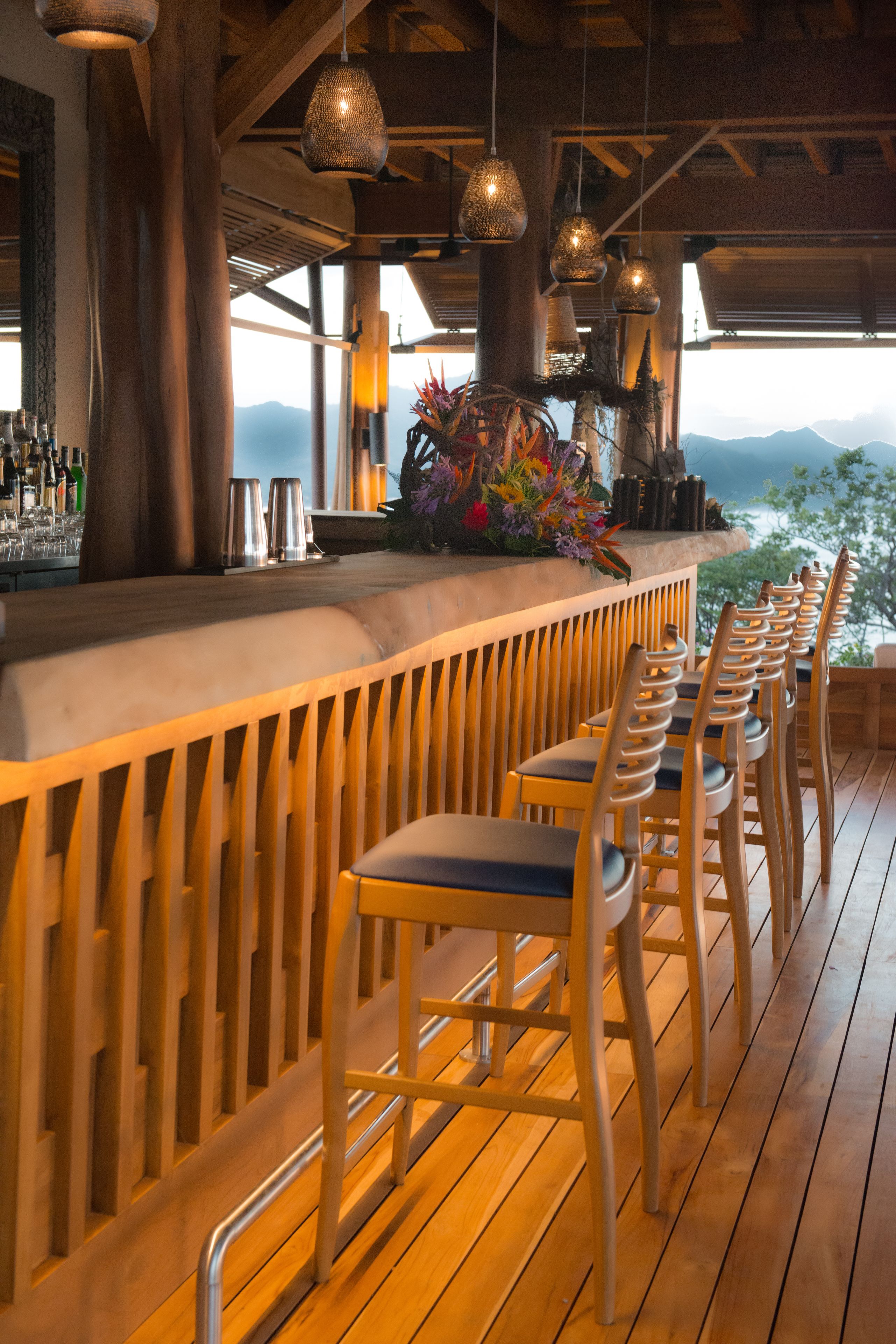 A bar area with wooden chairs and a bar counter, featuring hanging pendant lights and a view of mountains.