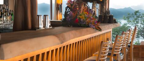A bar area with wooden chairs and a bar counter, featuring hanging pendant lights and a view of mountains.