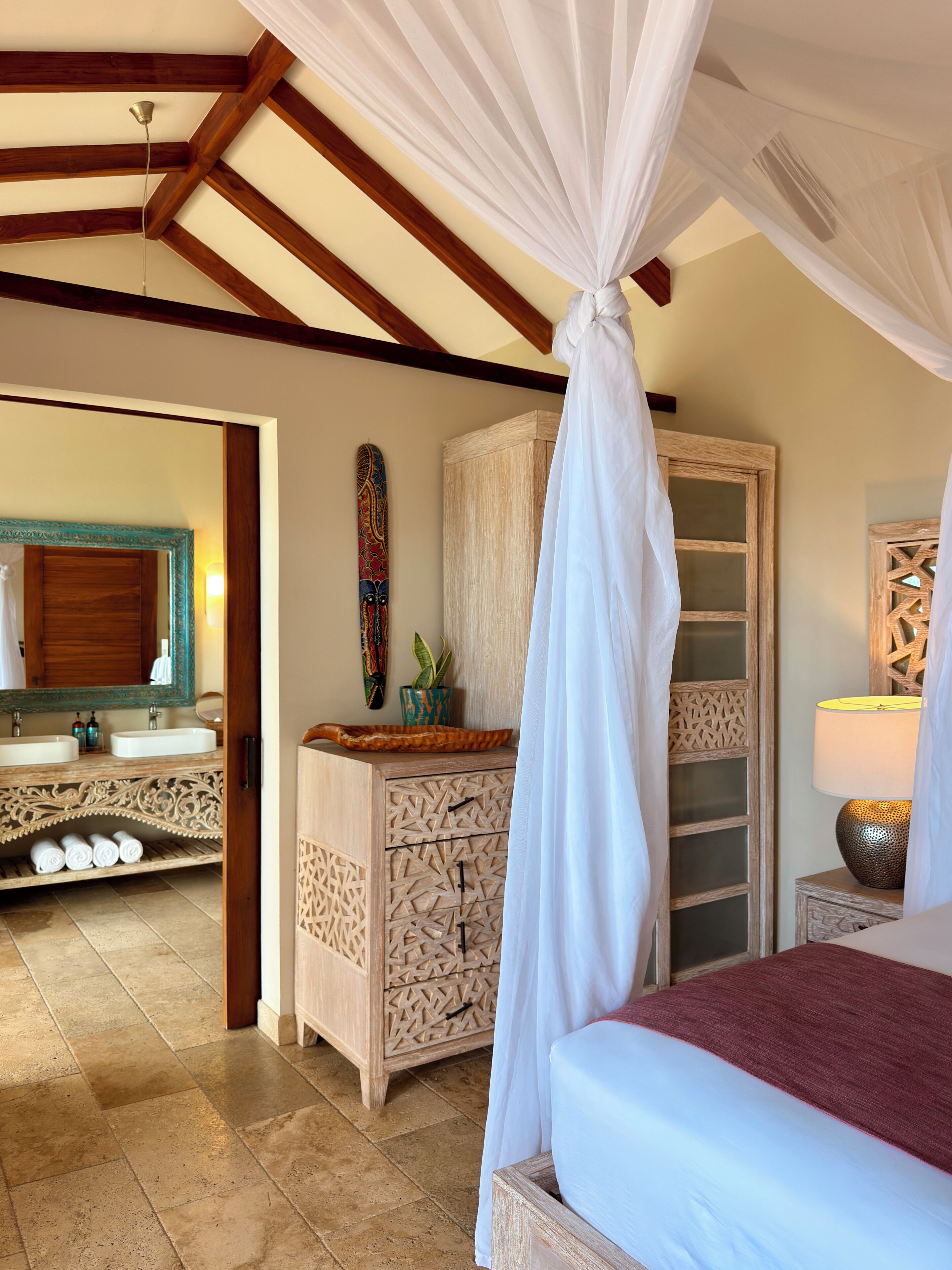 A bedroom with a canopy bed, a wooden cabinet with drawers, a mirror, and a bedside table with a lamp.