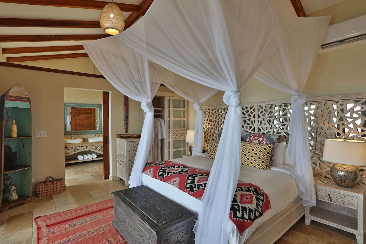 Un dormitorio con cama con dosel, una mesita de noche con lámpara, una mesilla auxiliar, un aparador y un panel decorativo en la pared.
