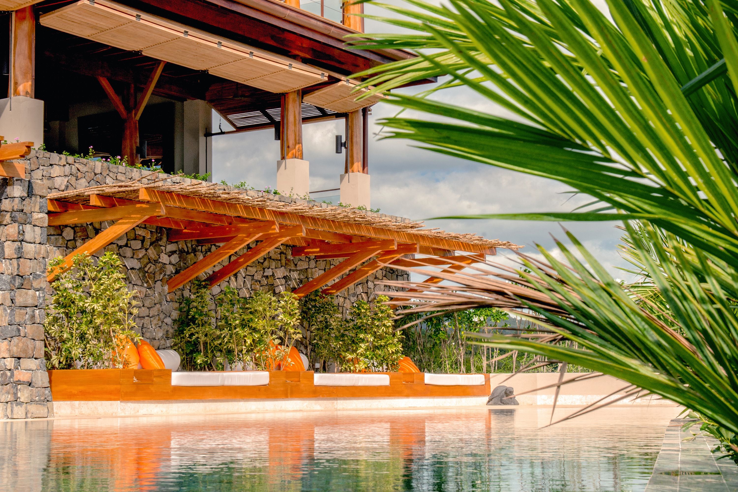 A pool area with a stone wall, wooden pergola, and tropical plants.