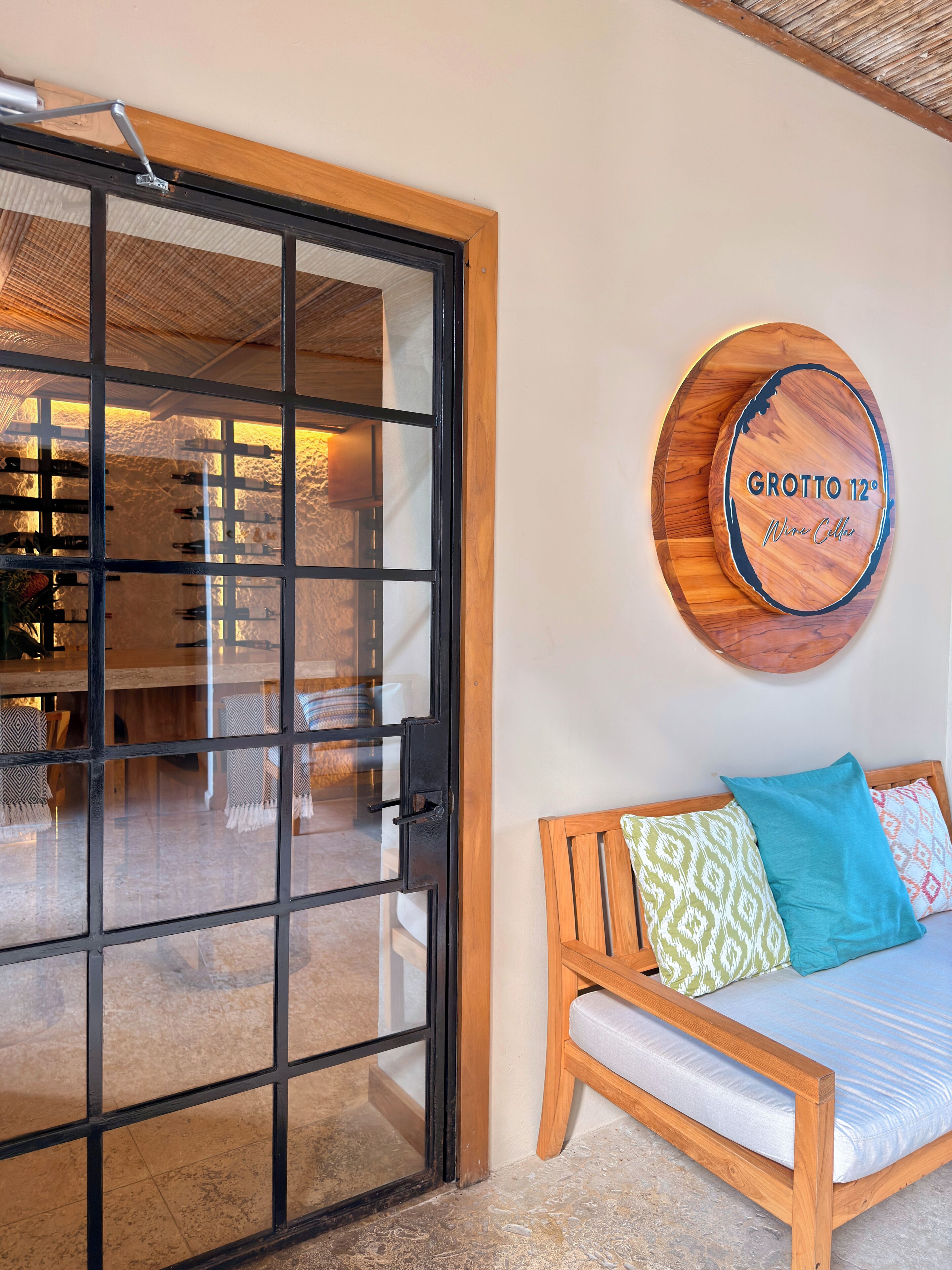 A room with a sliding glass door leading to a dining area, a round wooden sign with 'Grotto 12° Wine Cellar' text, and a wooden bed with patterned pillows.